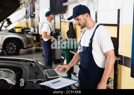 Selective focus of auto mechanic using laptop on car near clipboard at service station Stock Photo