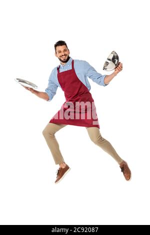 Positive waiter in apron holding tray and dish cover while jumping isolated on white Stock Photo