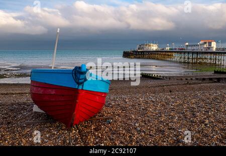 Worthing seafront West Sussex England UK - Red & blue fishing boat on the beach with Worthing Pier behind at dusk Stock Photo