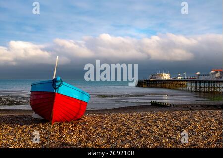 Worthing seafront West Sussex England UK - Red & blue fishing boat on the beach with Worthing Pier behind at dusk Stock Photo