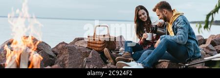 panoramic crop of happy woman pouring tea in cup near cheerful man and burning bonfire Stock Photo