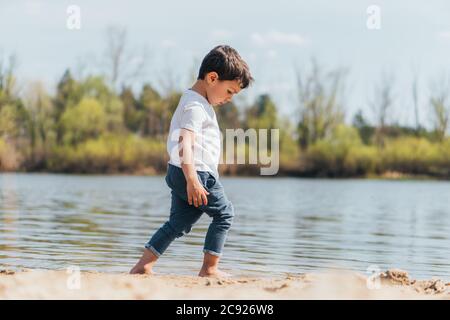 side view of barefoot boy walking on wet sand near pond Stock Photo