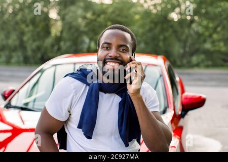 Positive handsome African American man standing in front of his luxury red car and looking at camera with happy smile, while having phone conversation Stock Photo