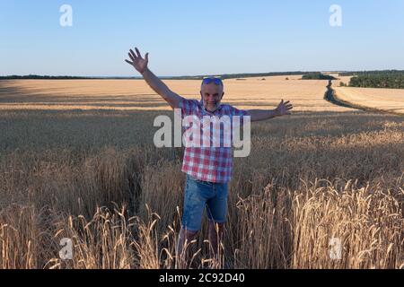A mature man stands in a wheat field. He spread his arms wide and smiles. Wearing a plaid shirt and shorts, sunglasses on the forehead. Stock Photo