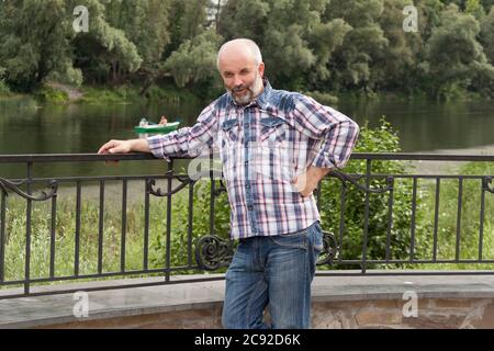 A man of mature age, bald with a beard, in a plaid shirt on the river embankment. It stands near the fence. Summer, abundant greenery, overcast Stock Photo