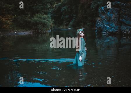 Young woman walking through the river in a green dress with flower wreath on her head Stock Photo