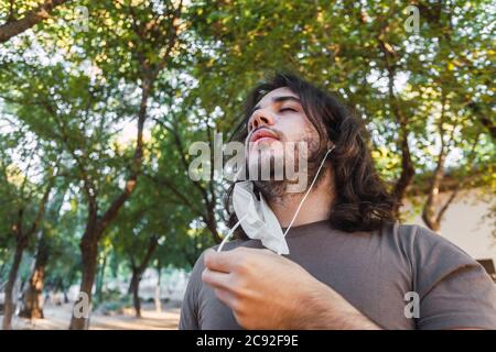 A young European man enjoying a breath of fresh air after removing face mask under lush trees Stock Photo