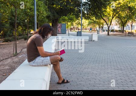 Male sitting on a stone bench in a park reading a book Stock Photo