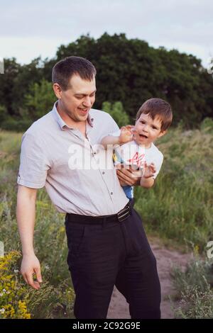 Dad holds his young son in his arms and hugs him in a summer field at sunset in the evening. Dad's love, attachment to the child. Stock Photo