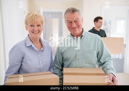 Portrait Of Senior Couple Downsizing In Retirement Carrying Boxes Into New Home On Moving Day With Removal Man Helping Stock Photo