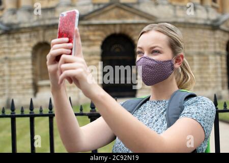 Female Tourist On Vacation Wearing Face Mask During Covid-19 Pandemic Taking Photo On Mobile Phone In Oxford UK Stock Photo