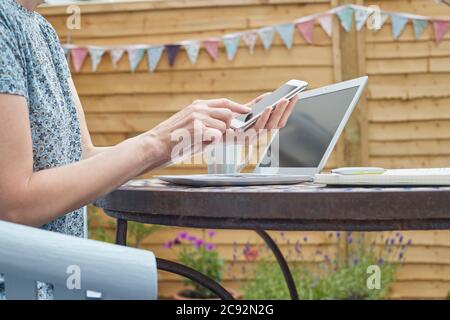 Woman Working From Home On Laptop Outdoors In Garden During Lockdown With Mobile Phone Stock Photo