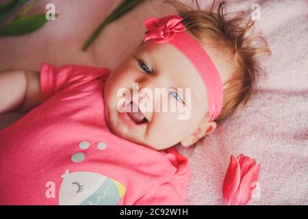Close-up portrait of cute happy smiling baby girl lying down on pink bed with tulips around. Stock Photo