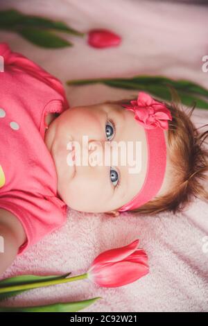 Close-up portrait of cute happy smiling baby girl lying down on pink bed with tulips around. Stock Photo