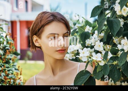 Attractive young girl with natural makeup in a white dress against the background of a blooming jasmine bush Stock Photo