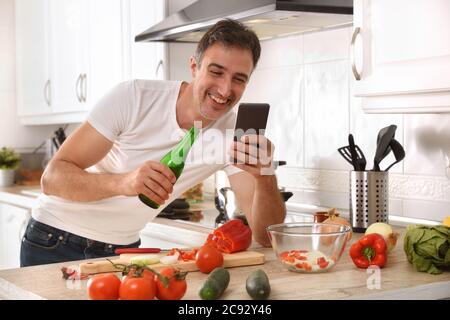 Happy man in the kitchen looking at a cell phone and having a beer while cooking leaning on the bench with vegetables Stock Photo