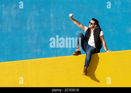 Young bearded man with sunglasses sitting on yellow wall,taking selfie Stock Photo