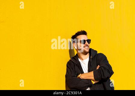 Young bearded man standing against a yellow wall with crossed arms Stock Photo