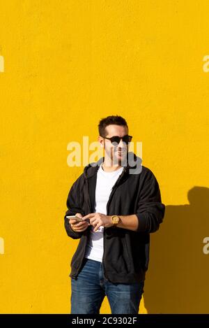 Bearded man with sunglasses standing against yellow wall, using phone Stock Photo