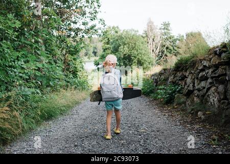 young girl walking down a country lane with a skateboard and backpack Stock Photo