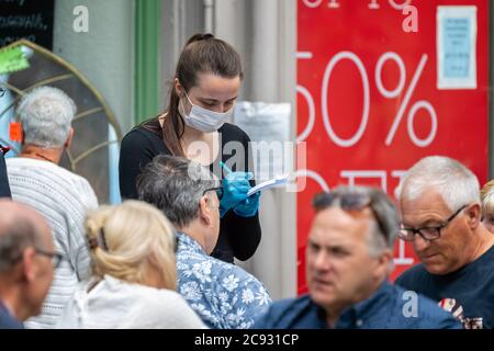 Young female waitress with mask and gloves taking coffee orders from customers outside in covid secure way - the new normal - in Wells, Somerset, UK o Stock Photo