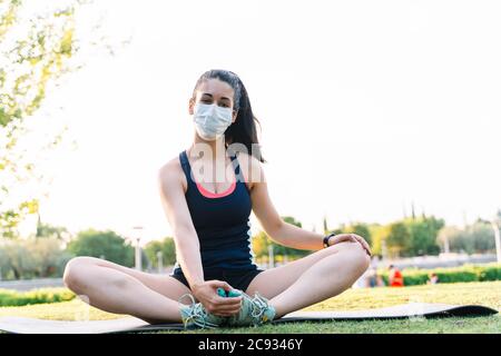 Woman with mask facing the camera while stretching on the ground in a public park. New normality concept Stock Photo