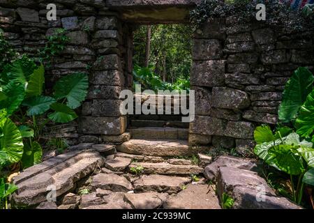 Entrance gate to the empty, ruined and overgrown by tropical plants ancient and old abandoned houses of a local village located in the middle of the l Stock Photo