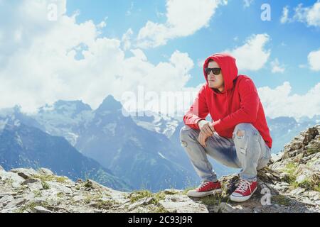 Young man in sunglasses sitting on mountain in sunny weather. Adult male in red hoodie with hood enjoying beautiful view in mountainous area. Stock Photo