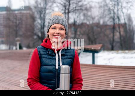 Beautiful Mature Woman Hot Holding Thermos In Park After Active Fitness Training With Friends. Active And Cheerful Lifestyle Of Middle Aged People Stock Photo
