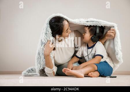 Asian mother hiding under blanket with infant child wearing headphones. Vietnamese mom smiling playing hide and seek with her son, family time concept Stock Photo