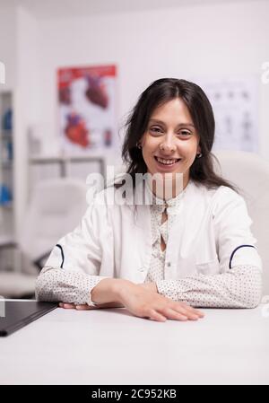 Portrait of smiling woman physician at desk in hospital office wearing white coat looking at the camera. Stock Photo