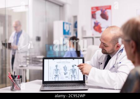 Doctor pointing at human skeleton on laptop in hospital office during consultation of old woman before surgery. Senior medic wearing white coat taking notes on clipboard in clinic corridor. Stock Photo