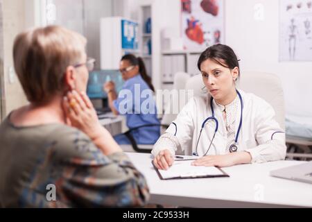 Young doctor examining senior patient in hospital cabinet wearing white coat and stethoscope. Mature woman with sore neck. Nurse working on computer. Stock Photo