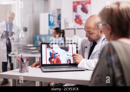 Cardiologist in white coat pointing at heart on laptop screen in hospital office and talking with old woman about treatment for her illness. Mature doctor writing prescription on clipboard in clinic corridor. Stock Photo