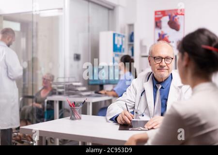 Senior doctor consulting woman patient in hospital office while younger colleague is discussing with disabled elderly aged woman in wheelchair on clinic corridor and nurse in blue uniform looking at x-ray on computer screen. Stock Photo