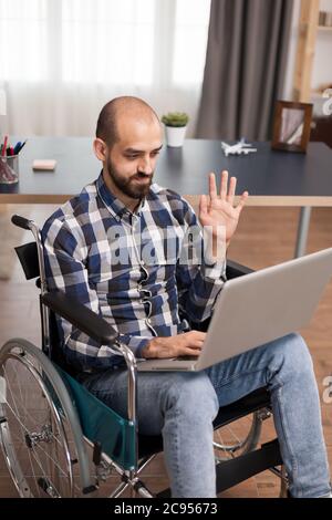 Disabled freelancer in wheelchair during a video call working from home. Businessman waving during video conference, working remotely from home on laptop. Stock Photo