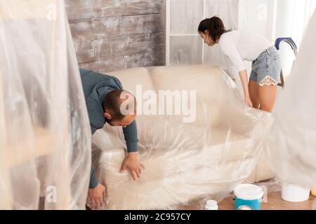 Couple wrapping sofa in plastic foil for protection while they are renovate living room. Home during renovation, decoration and painting. Interior apartment improvement maintenance. Roller, ladder for house repair Stock Photo