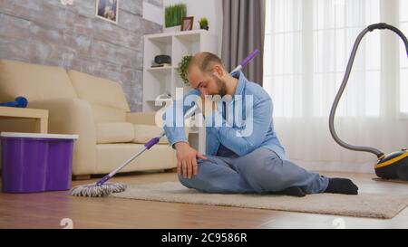 Tired man sitting on cozy carpet after cleaning the room floor with mop and detergent. Stock Photo