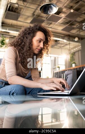 Focused curly-haired young woman immersed into social media typing on laptop while interacting online in cafe Stock Photo