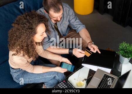 Young male cafe owner with beard sitting on sofa and pointing at tablet screen while talking to waiter about menu Stock Photo