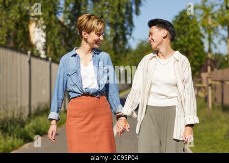 Two young happy girlfriends smiling to each other and holding hands while walking in the park Stock Photo