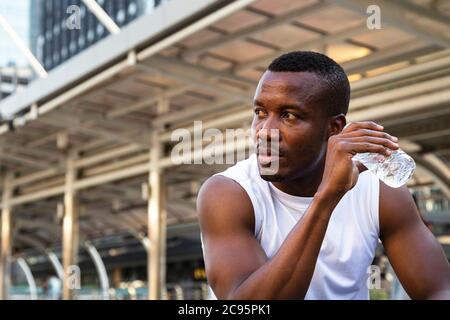 portrait of relaxing young african american sportsman sitting and drinking water from bottle after running exercise in city in morning with building b