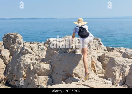 Woman traveler wearing straw summer hat and backpack, climbing at edge of the rocky cliff and looking at big blue sea and islands in on the horizon Stock Photo