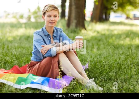 Portrait of young woman with short blonde hair smiling at camera while sitting on the grass and drinking coffee in the park Stock Photo
