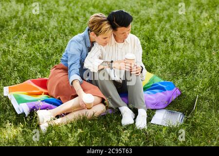 Two girlfriends sitting on the grass drinking coffee and watching something on laptop computer during picnic outdoors Stock Photo