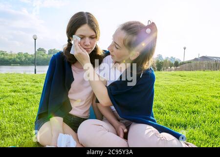 Crying teenager girl and comforting girlfriend, teenagers sitting on the grass in park Stock Photo