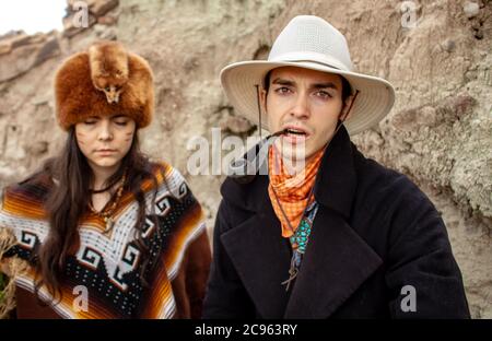 A trapper woman with a poncho and fur hat and serious cowboy wearing a hat, smoking a tobacco pipe in desert canyon in Ojito Wilderness, New Mexico Stock Photo