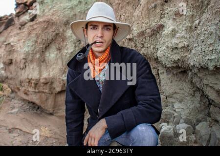Serious cowboy wearing a wide brimmed hat and bandanna smokes a tobacco pipe in an arid desert canyon in the Ojito Wilderness, New Mexico Stock Photo