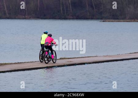 2 cyclists cycling & riding bikes (cycle ride) on narrow causeway pathway - scenic rural RSPB nature reserve lake, Leeds, West Yorkshire, England, UK. Stock Photo
