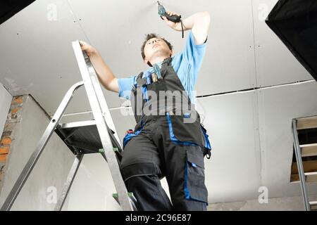 Guy in work clothes and with an electric screwdriver in his hand is standing on the stairs. Against the background of renovation in the house Stock Photo